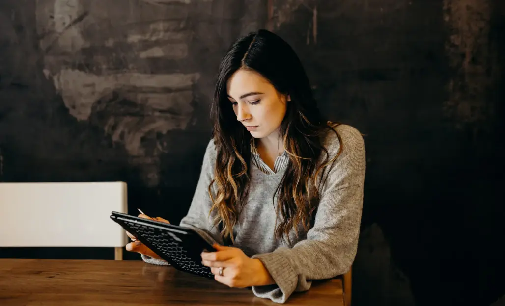 Een vrouw zit aan een houten tafel en is geconcentreerd bezig met een tablet in een donkere, warme omgeving.