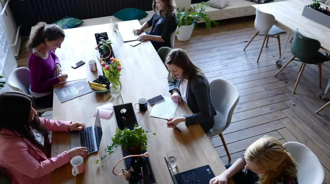 Een groep mensen die samenwerkt rond een grote houten tafel, met computers, planten en bekers, in een lichte en open werkomgeving.